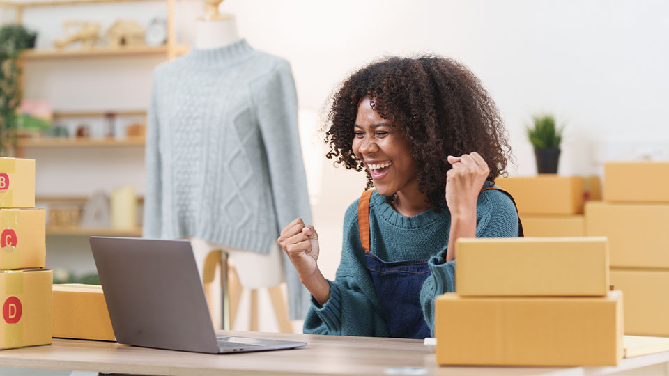 A woman with curly hair is sitting at a desk surrounded by shipping boxes and a laptop. She looks excited, raising her fists triumphantly. In the background, there's a mannequin wearing a sweater and some shelves with decor.