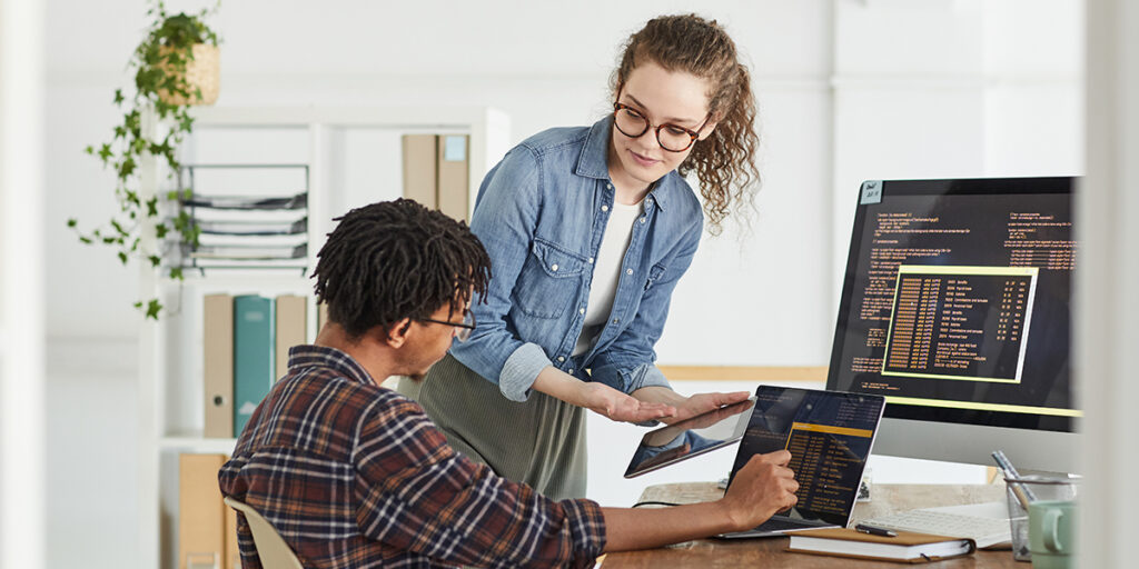 A woman shows a tablet to a man seated at a desk with a laptop. Both are focused on the screen displaying code. A large monitor with code and an organized shelf with plants are visible in the bright office.
