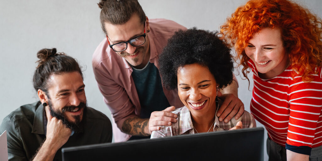 A diverse group of four people smiling and looking at a computer screen. They appear to be in a positive and collaborative atmosphere, with one person pointing and others leaning in closely. The setting looks like an office or workspace.