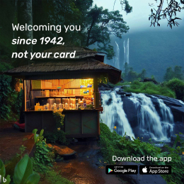 A cozy, illuminated wooden shop nestled beside a lush waterfall in a scenic forest setting, with the text 'Welcoming you since 1942, not your card' displayed. App store icons for Google Play and the App Store are shown, encouraging app downloads.