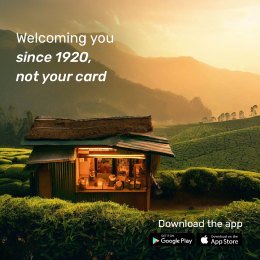 A small, warmly lit shop situated in the middle of a lush green tea plantation at sunset, with the text 'Welcoming you since 1920, not your card' displayed. App store icons for Google Play and the App Store are shown, promoting app downloads.