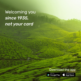 A vast, vibrant green tea plantation stretching across rolling hills under soft sunlight, with the text 'Welcoming you since 1935, not your card' displayed. App store icons for Google Play and the App Store are shown, encouraging app downloads.