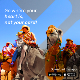 A group of people riding camels in a desert, wearing colorful clothing and sun hats. The text reads "Go where your heart is, not your card!" An advertisement for a travel app, with download icons for Google Play and App Store.