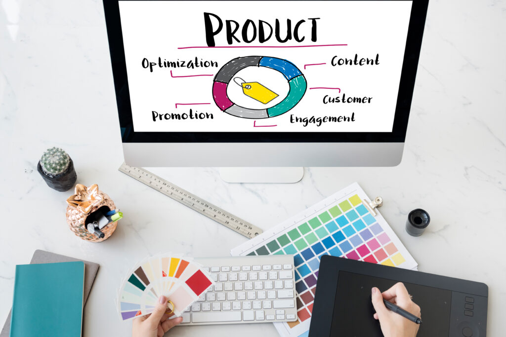 Computer screen displaying a product strategy diagram, surrounded by a keyboard, color swatches, a digital drawing tablet, a ruler, a small plant, and a decorative jar on a desk.