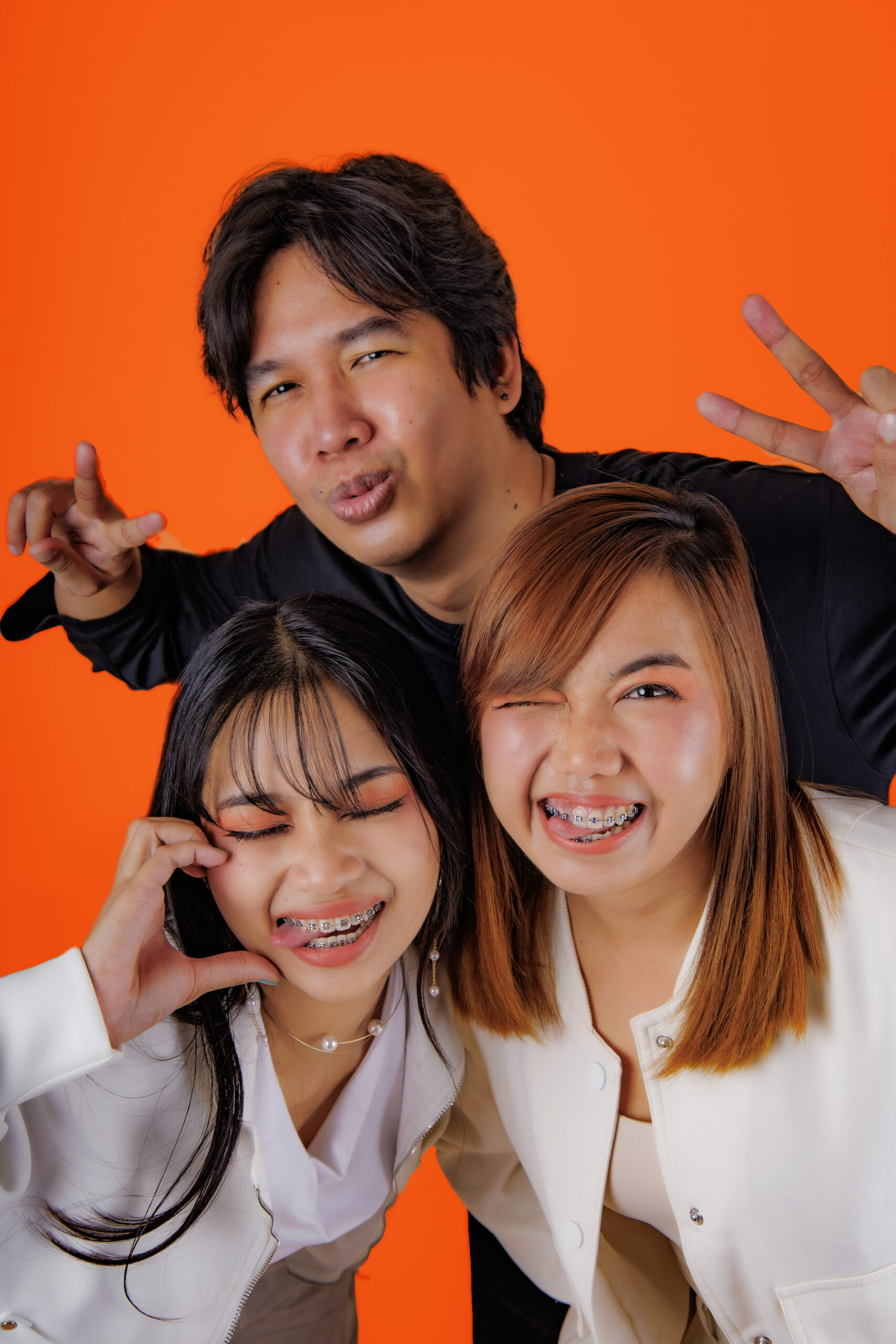 Three young people with braces pose for a playful photo against an orange background, making peace signs and funny faces.