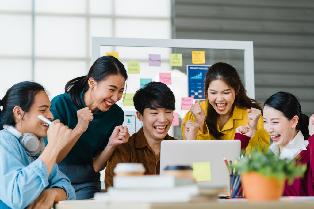 A group of five diverse young adults excitedly react to a laptop screen in a bright office. A whiteboard with colorful sticky notes is in the background.