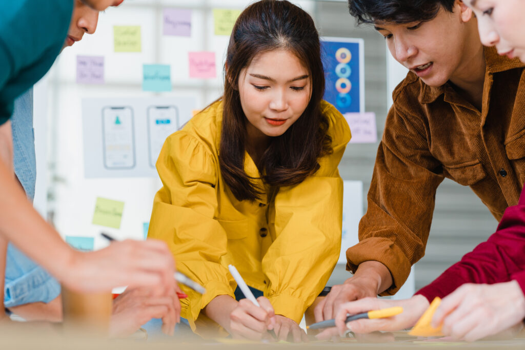 A group of young adults collaborates intensely around a table, writing with markers. The scene is dynamic and focused, with colorful sticky notes in the background.
