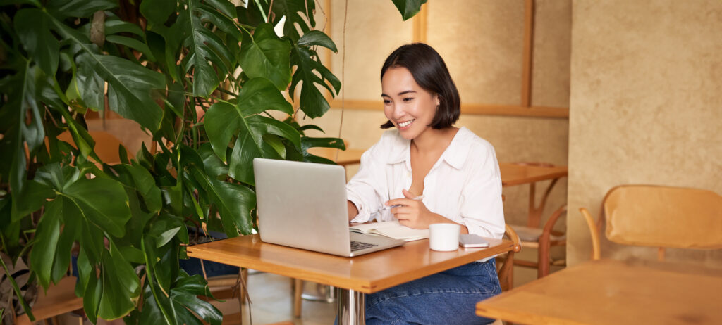 Woman sitting at a table in a cafe, using a laptop with a notebook and cup in front of her, smiling while working. Large green plant beside her.